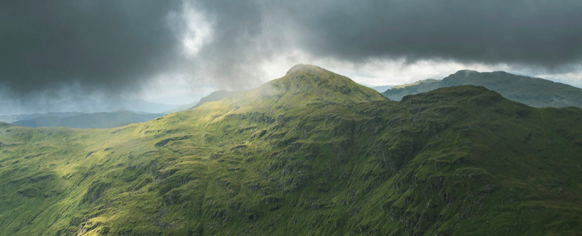 Lande de montagnes vertes sous un ciel dramatique, dans l'esprit des Highlands écossais de Skyfall — l'univers de Daniel Craig et de Grey Vetiver de Tom Ford, par la parfumerie Olara